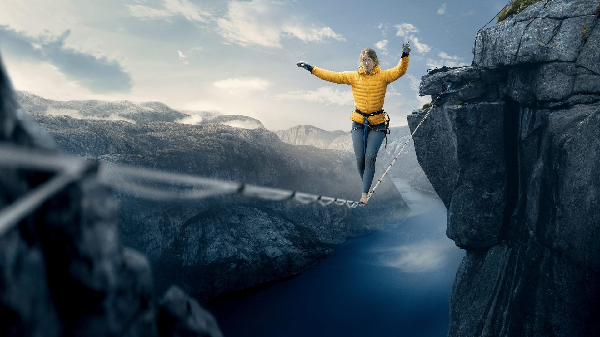 A woman in blue leggings and a yellow puffy jacket crosses a slack line between two cliffs above a Norwegian fjord.