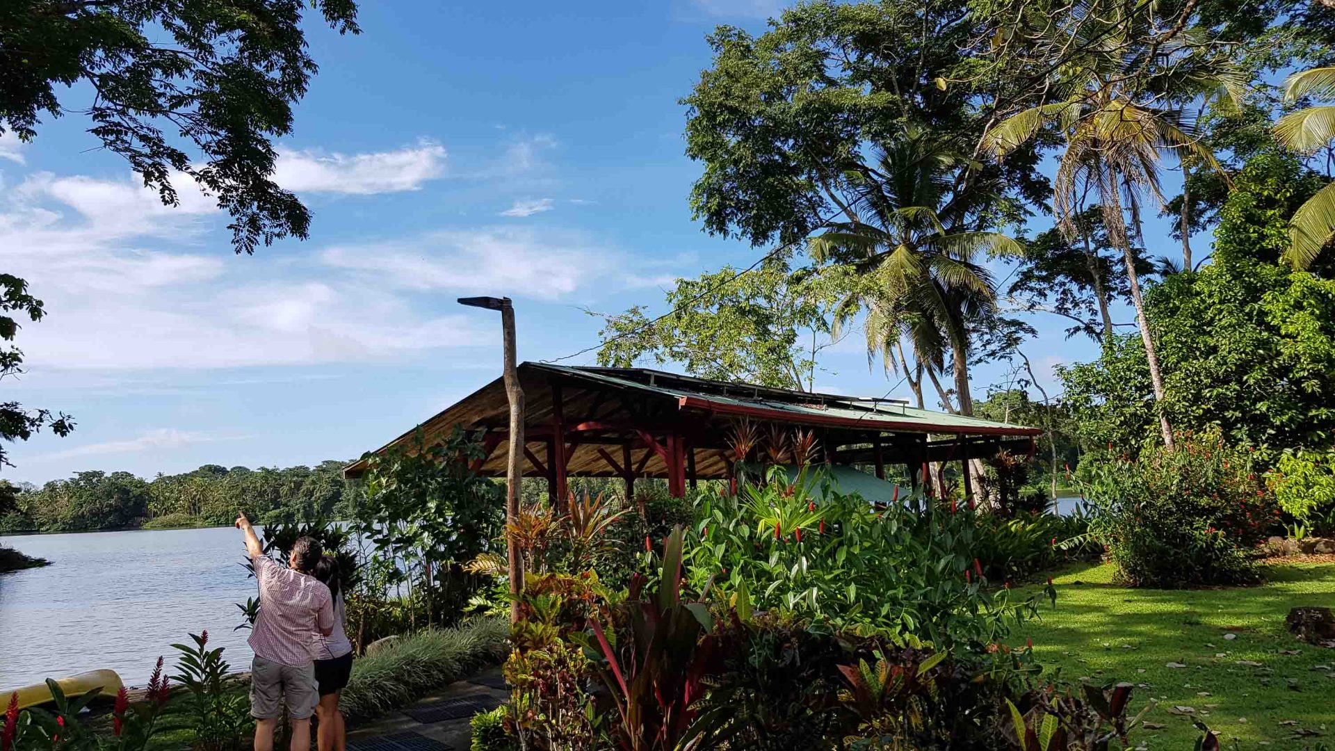 Two people point to the trees while standing alongside a lodge on the river.