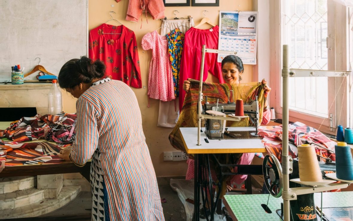 The interior of a sewing workshop. At the back, a woman smiles and holds up a piece of colorful fabric while sat at her sewing machine, while another woman sorts through fabrics at a table.