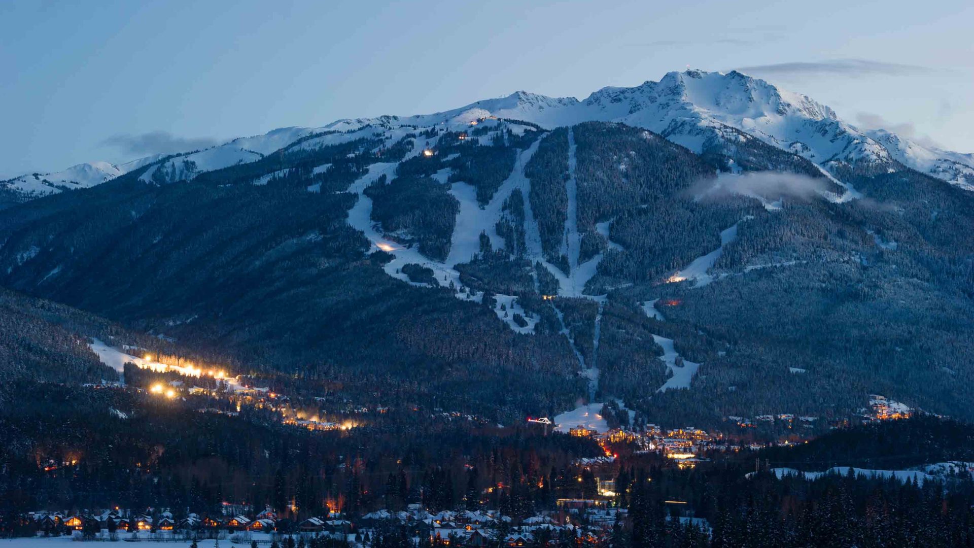 Ski fields on a mountain descend to a town lit up at dusk.