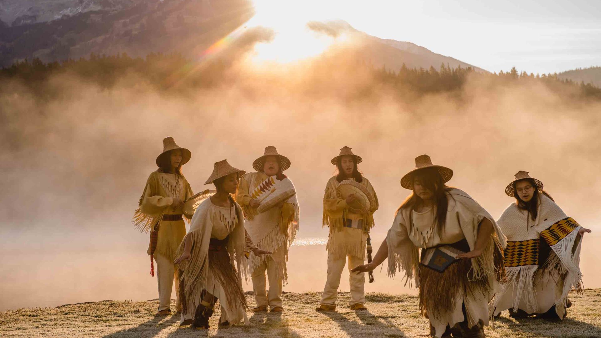 First Nations people perform a ceremony in soft light in front of some mountains.