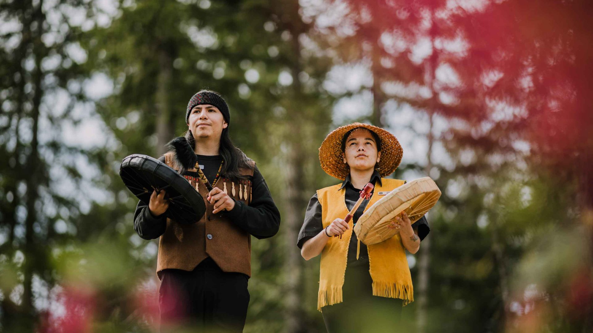 Two First Nations people hold musical instruments and look up to the trees.