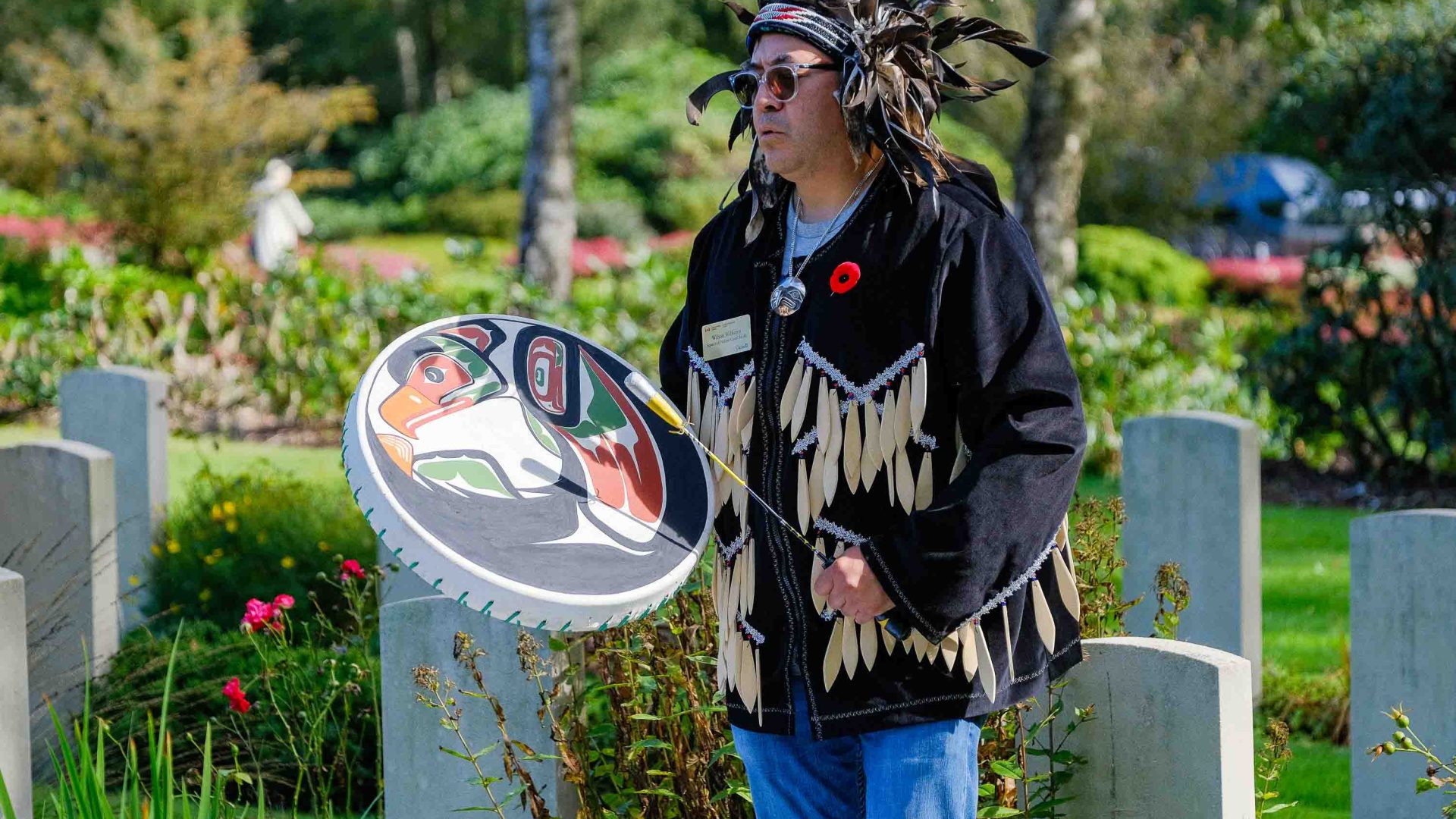 A man in a traditional head dress and clothing stands outside alongside a drum.