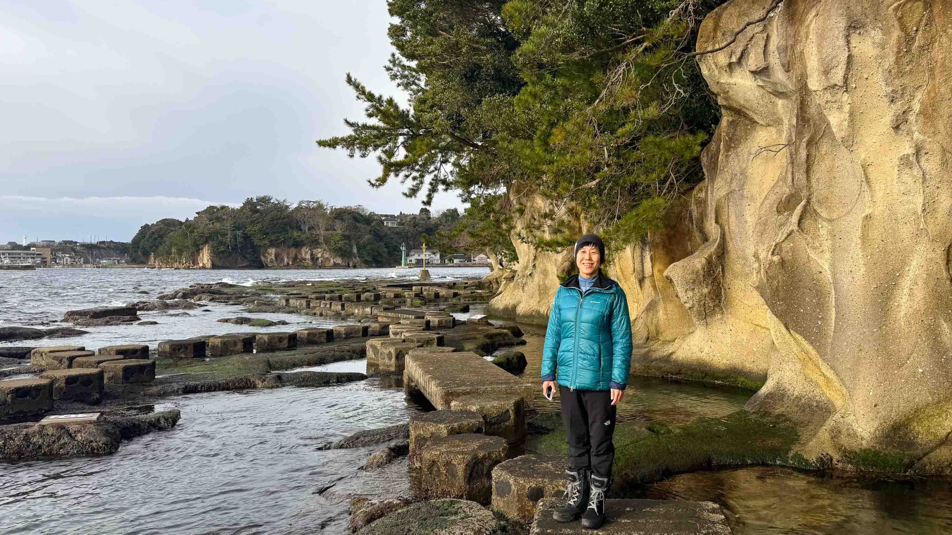 A woman stands and smiles to camera while standing on rocks in a bay.