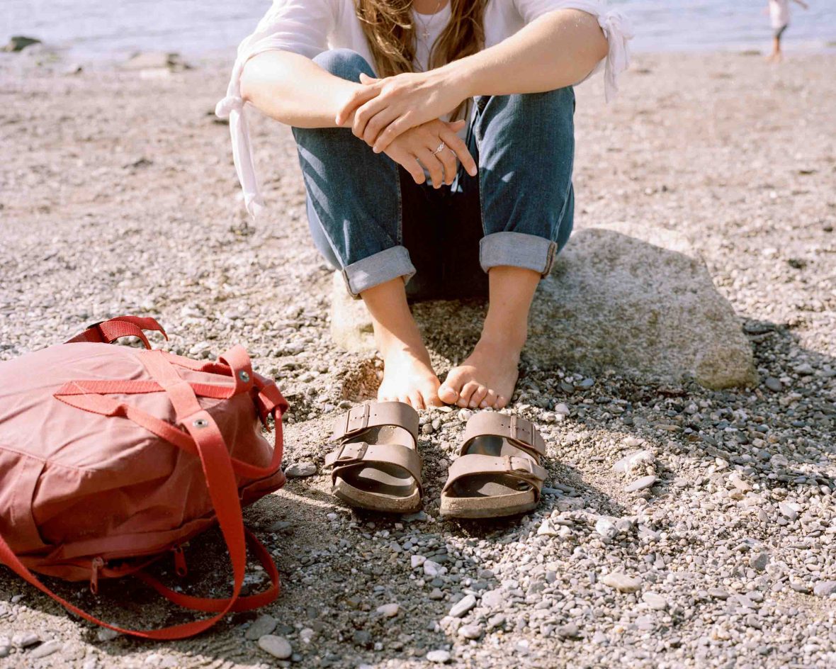 A woman sits in her bare feet, with her Birkenstock sandals in front of her on the sand of a beach.