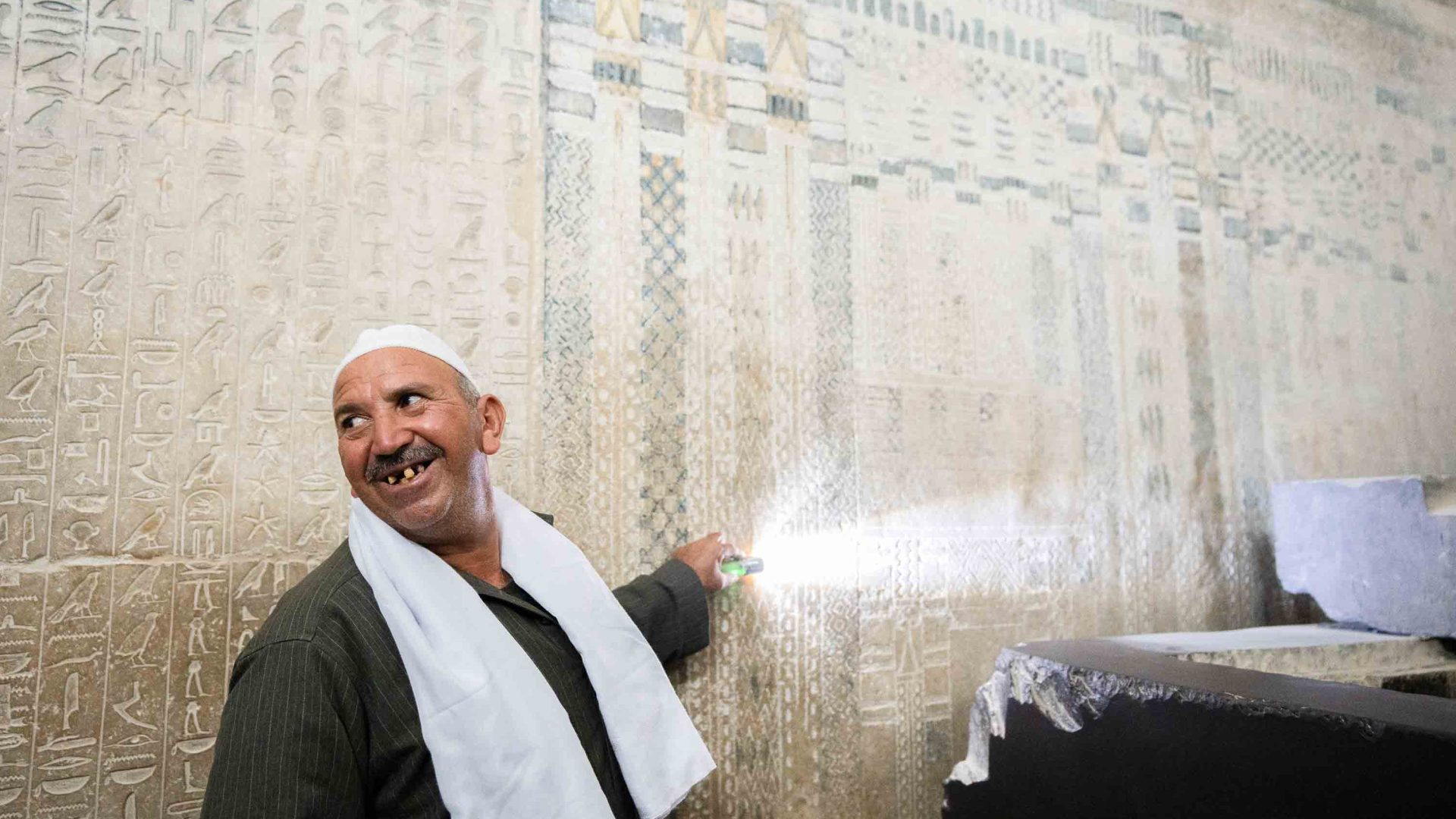 A man smiles as he shines a light on a wall with hieroglyphics on it.