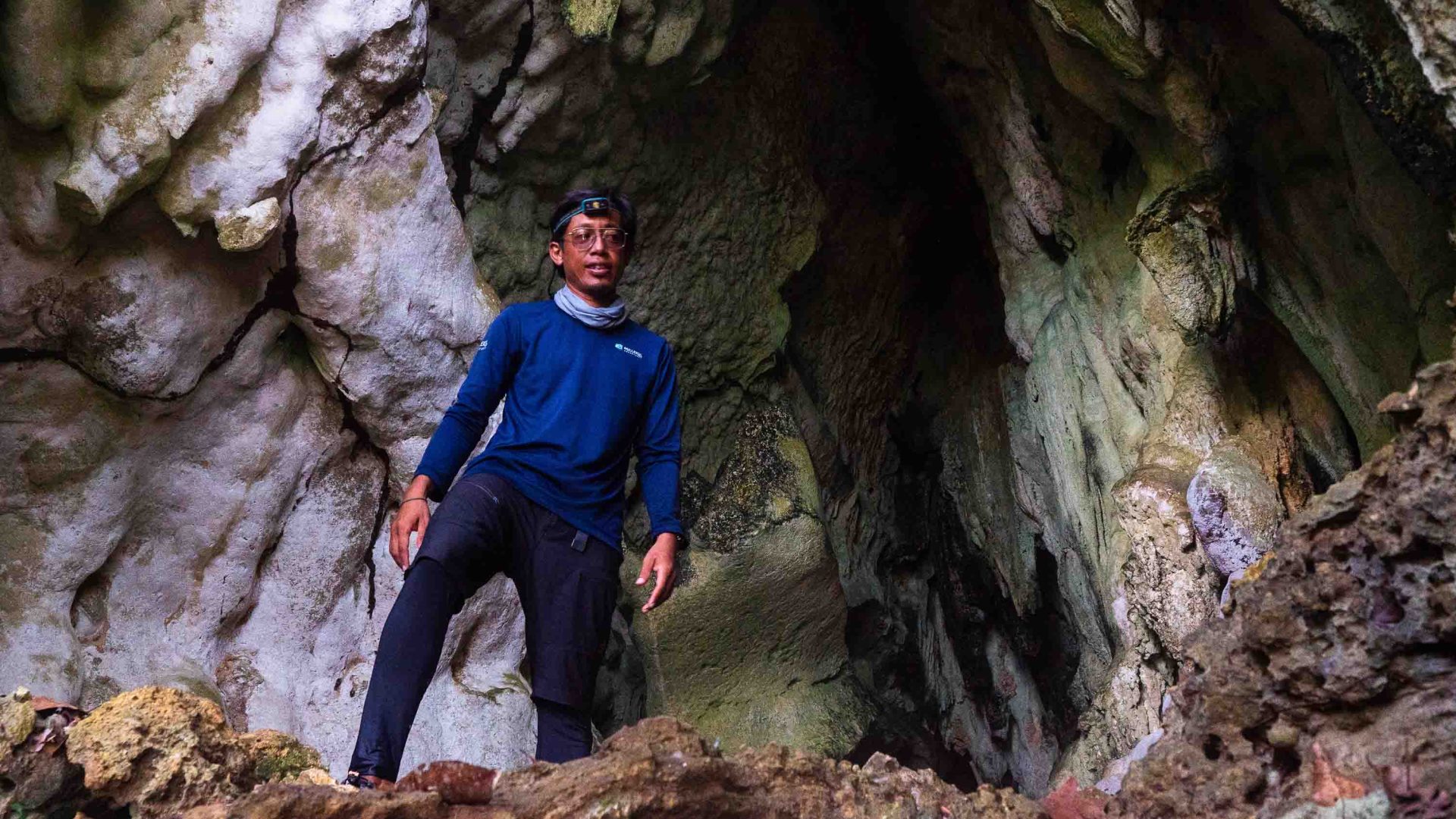 A man stands at the mouth of a cave wearing a head torch.