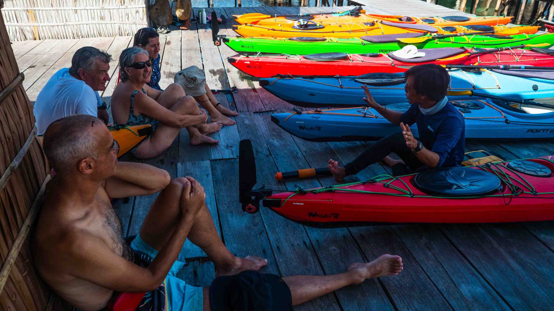 A group of people sit and listen to a man instruct them on kayaking. They have kayaks next to them.