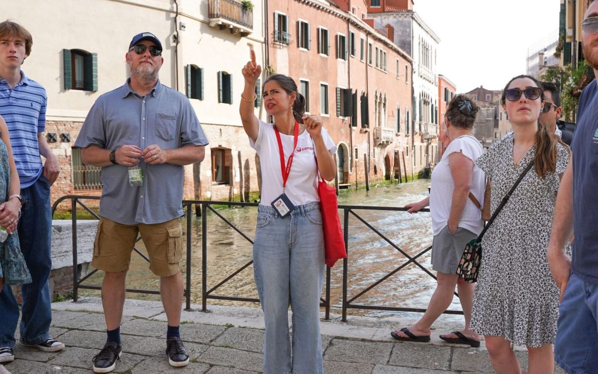 A woman stands on a bridge over a Venice canal as she guides a group around the city