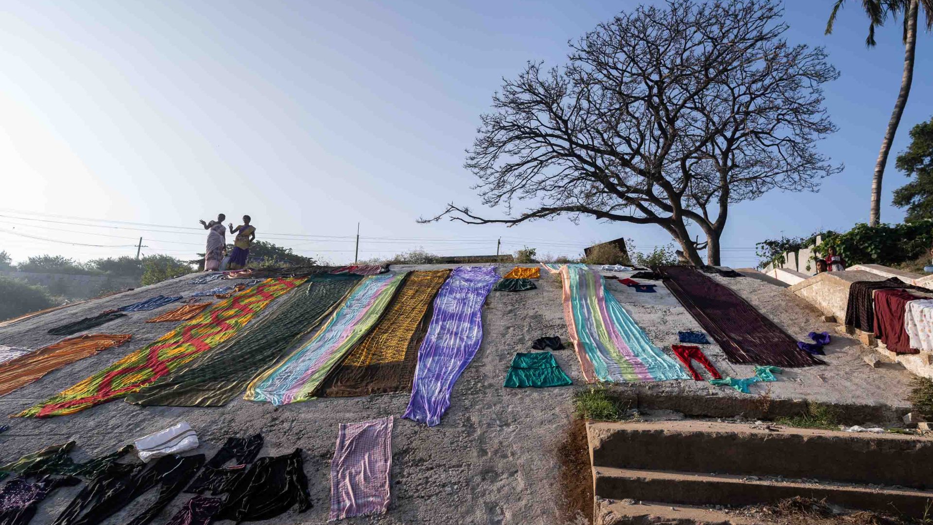 Two people stand on a hill where rows of colooured cloth dry in the sun.