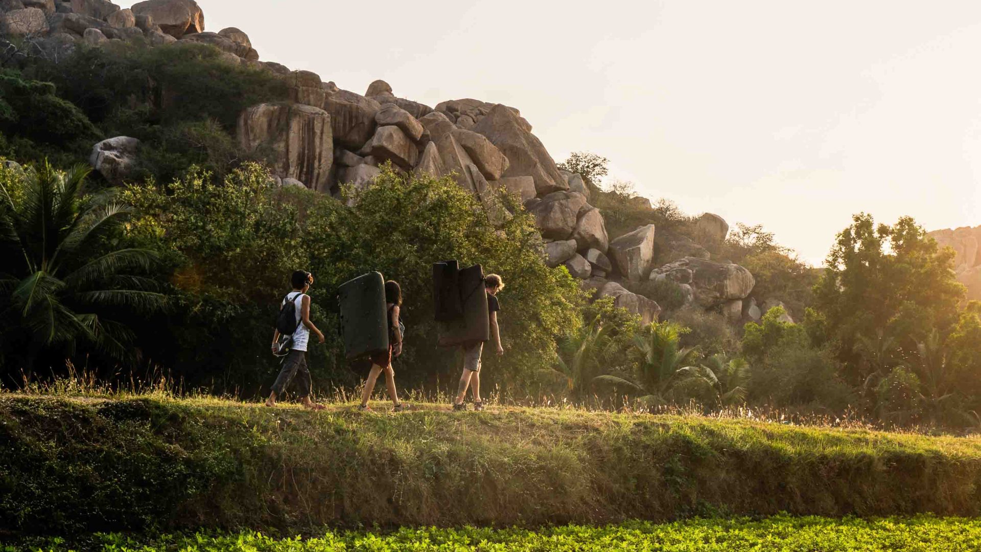 A group of people carry crash pads through green fields with boulders in the background.