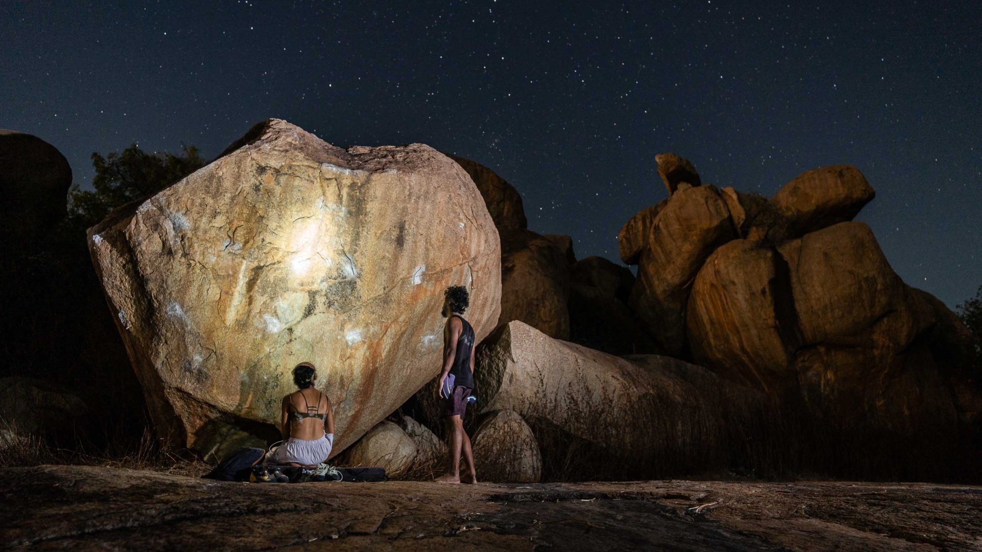 Two people look at a boulder at night and appear to be having a conversation.