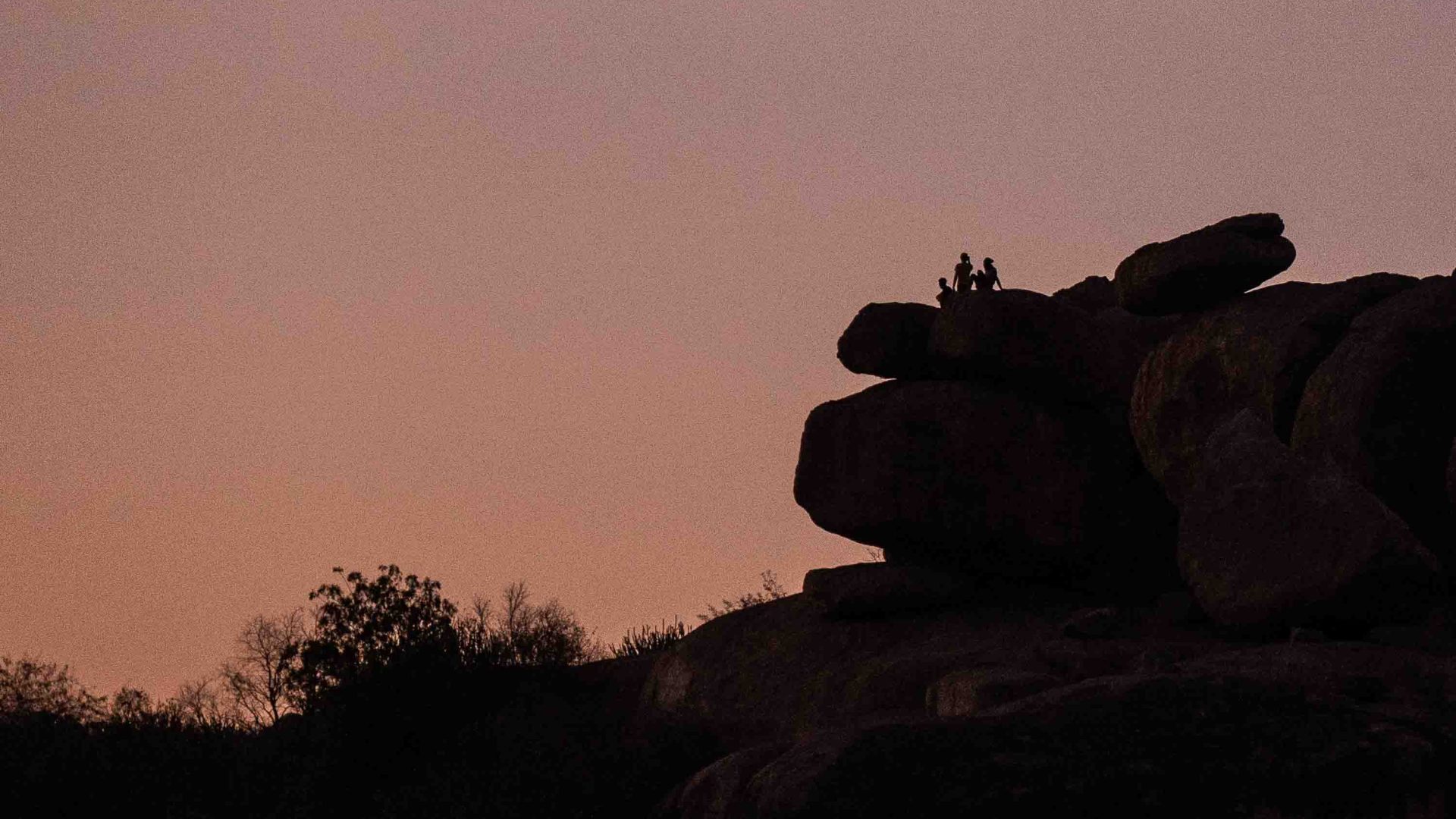 Three people sit on the edge of a rock and watch a sunset.