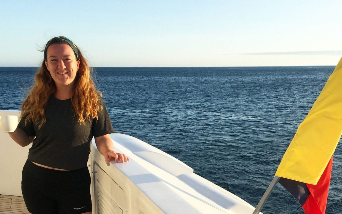 A woman smiles to camera holding a cup of tea by the side of a ship at sea