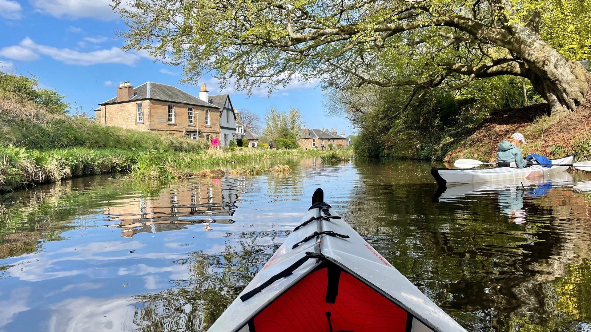 A spring day on a canal in Scotland, with a historic home in the background and a tree extending over the canal in the foreground. The photographer is sitting inside a kayak and the front of the boat is visible. Another woman in a kayak is off to the right.