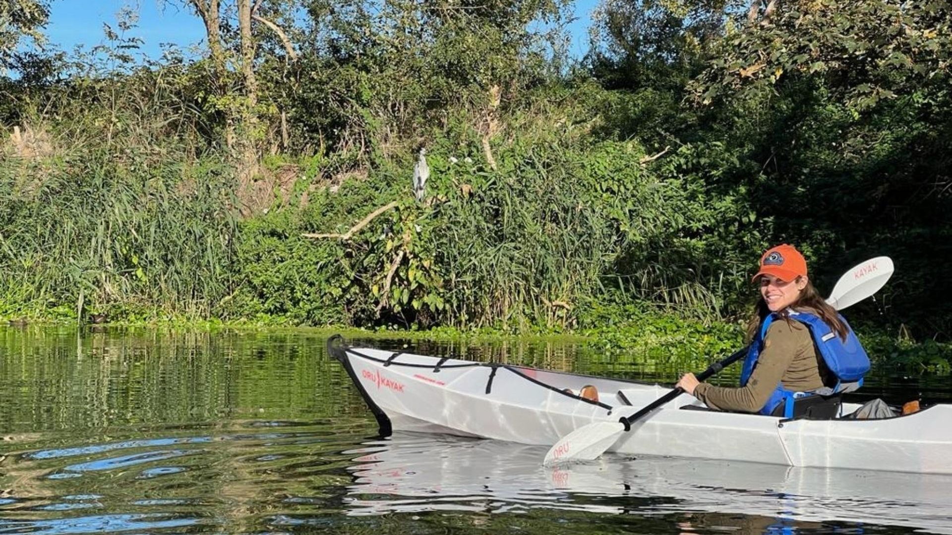 A woman in a white kayak smiles toward the camera. A heron is visible in the background, among wild brush.