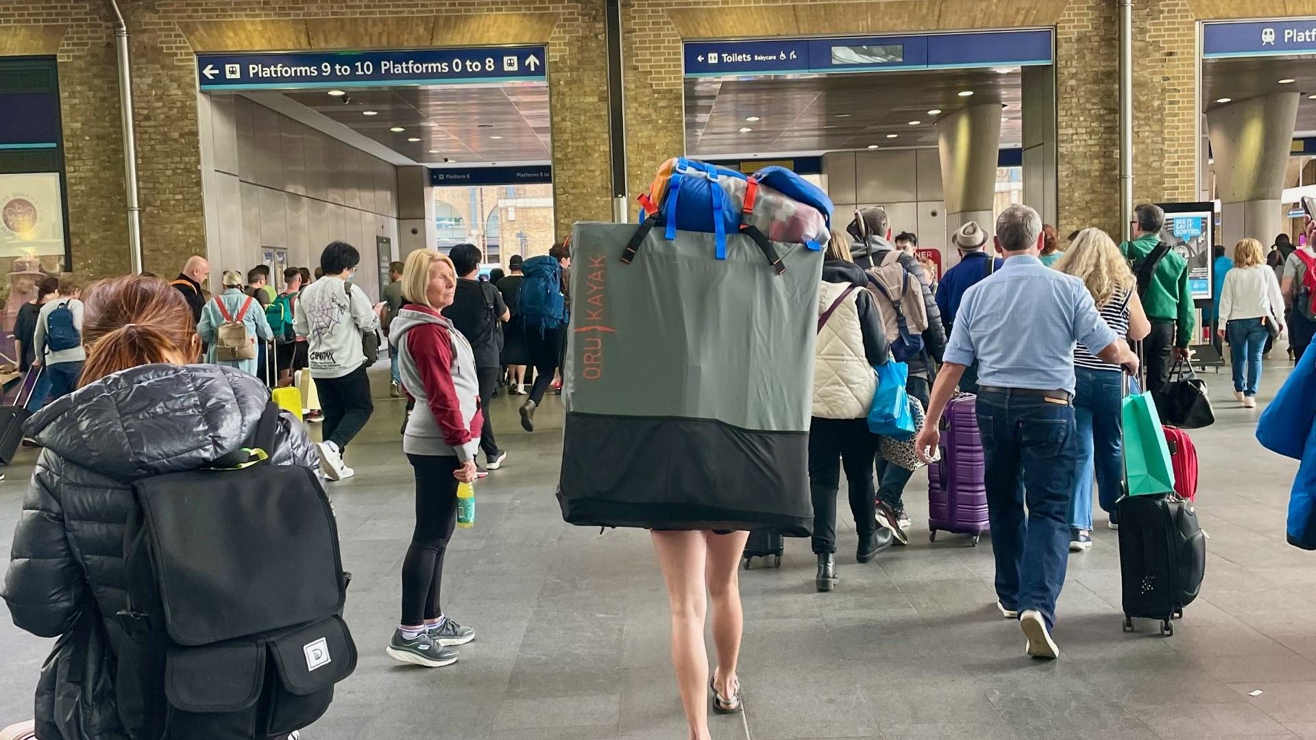 A woman carries a large, square backpack through Kings Cross train station. Only her legs are visible. It's a busy day.