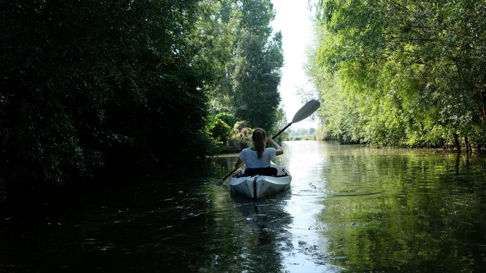 A woman paddles a white kayak through a tunnel of trees in Saint-Omer, France.