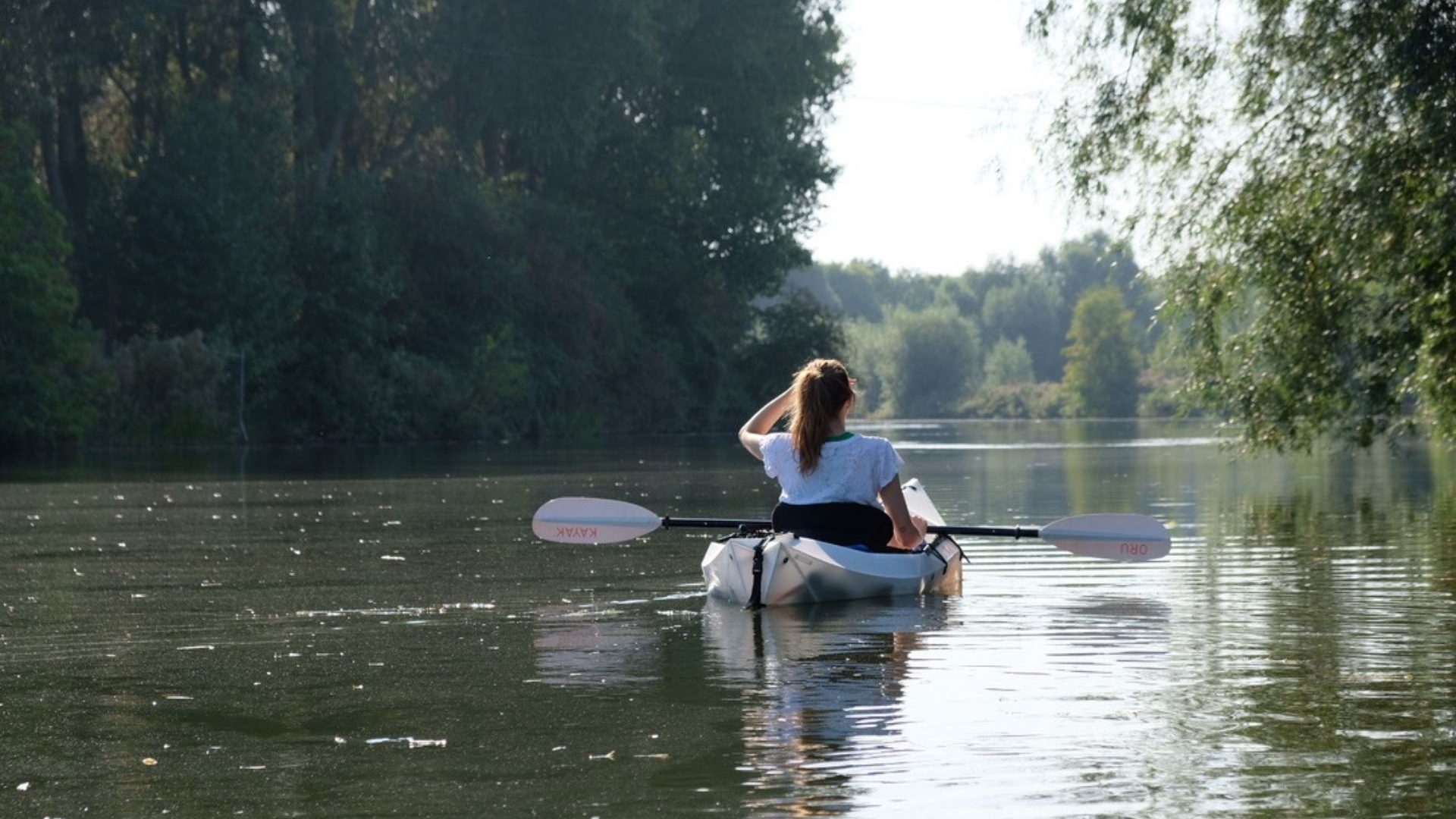 A woman in a white kayak faces away from the camera, with her hand up to shade her eyes from the sun. The canal is green and lined with trees on both sides.