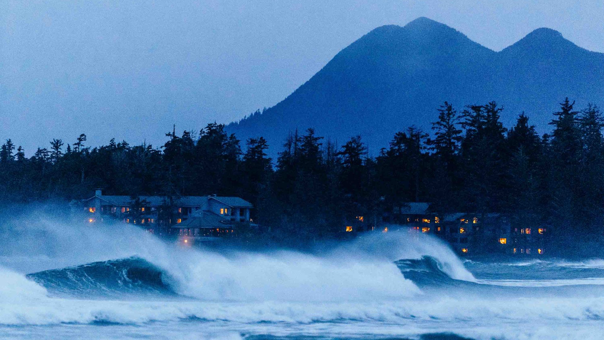 The Inn is lit up at night as waves crash in front of it and a mountain stands behind it.