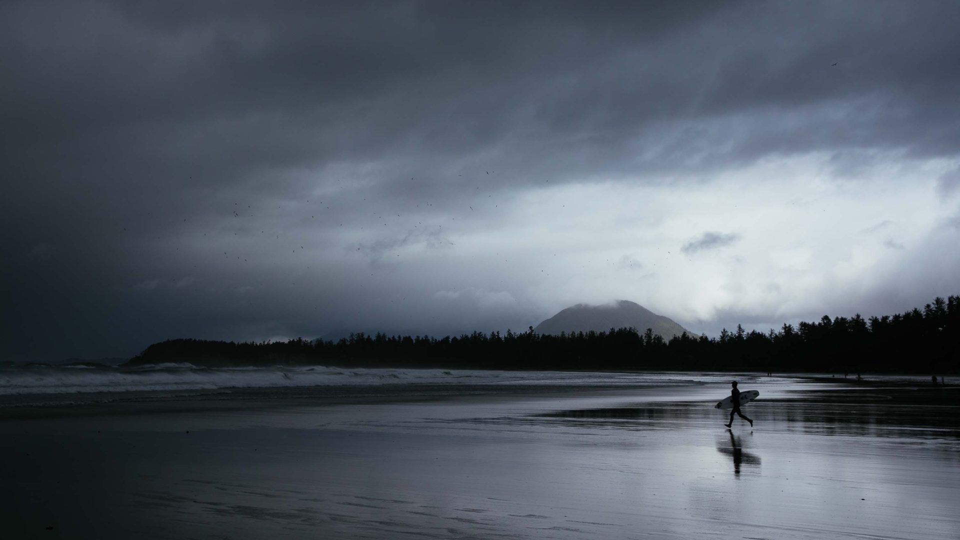 A person walks toward the sea with his surfboard. The sky is grey and cloudy.