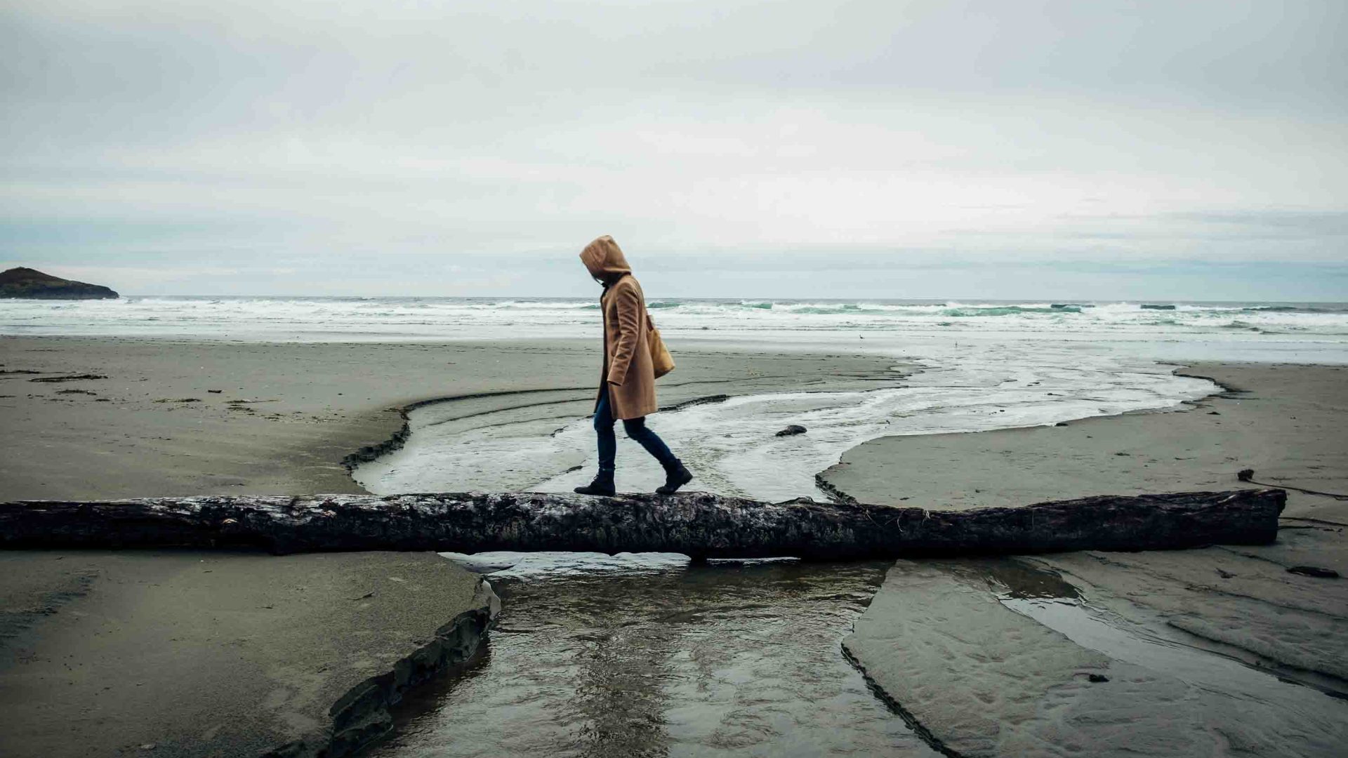 A person crosses over some driftwood on a beach.