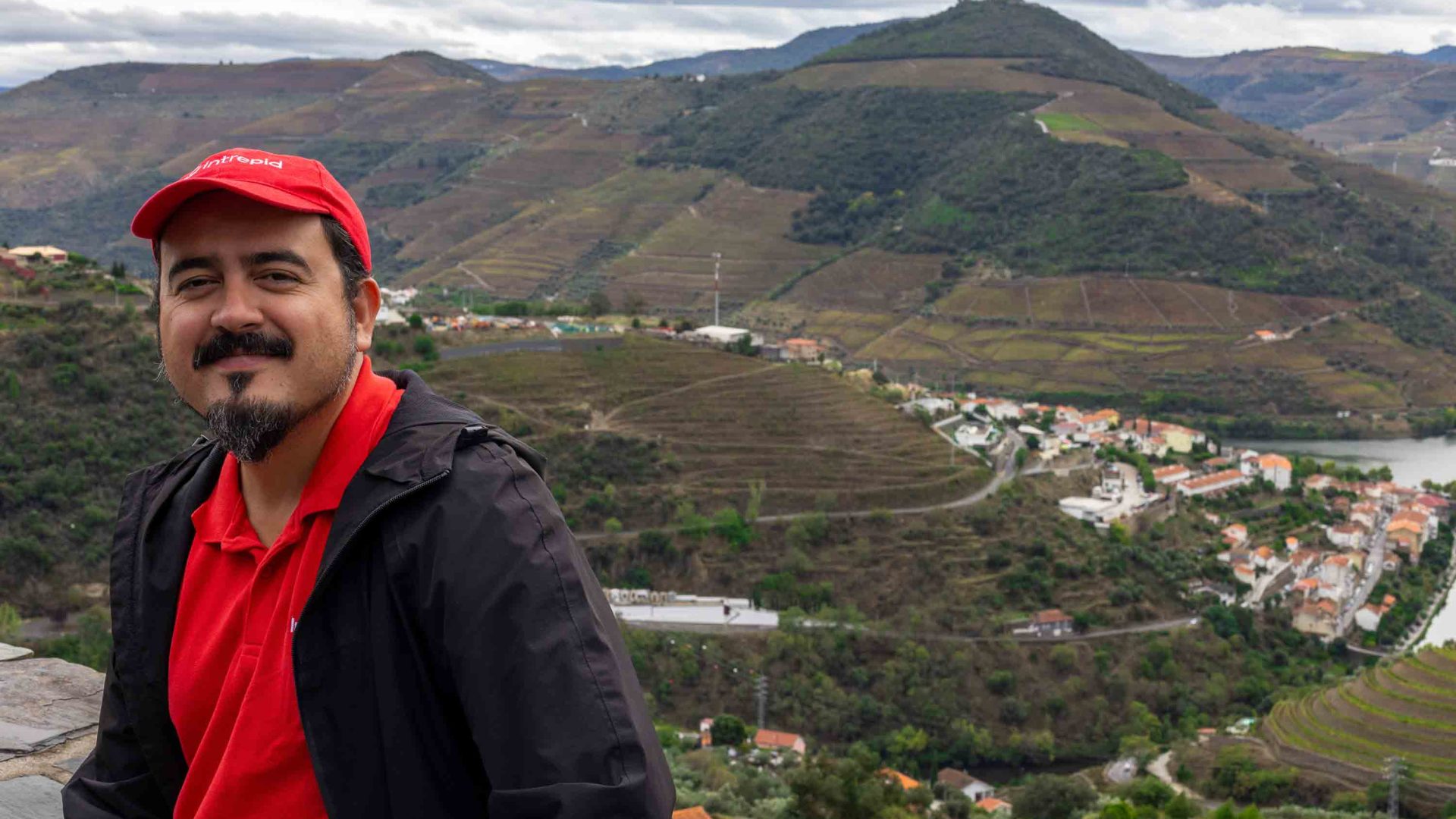 A man in red smiles as he stands above a valley with river.