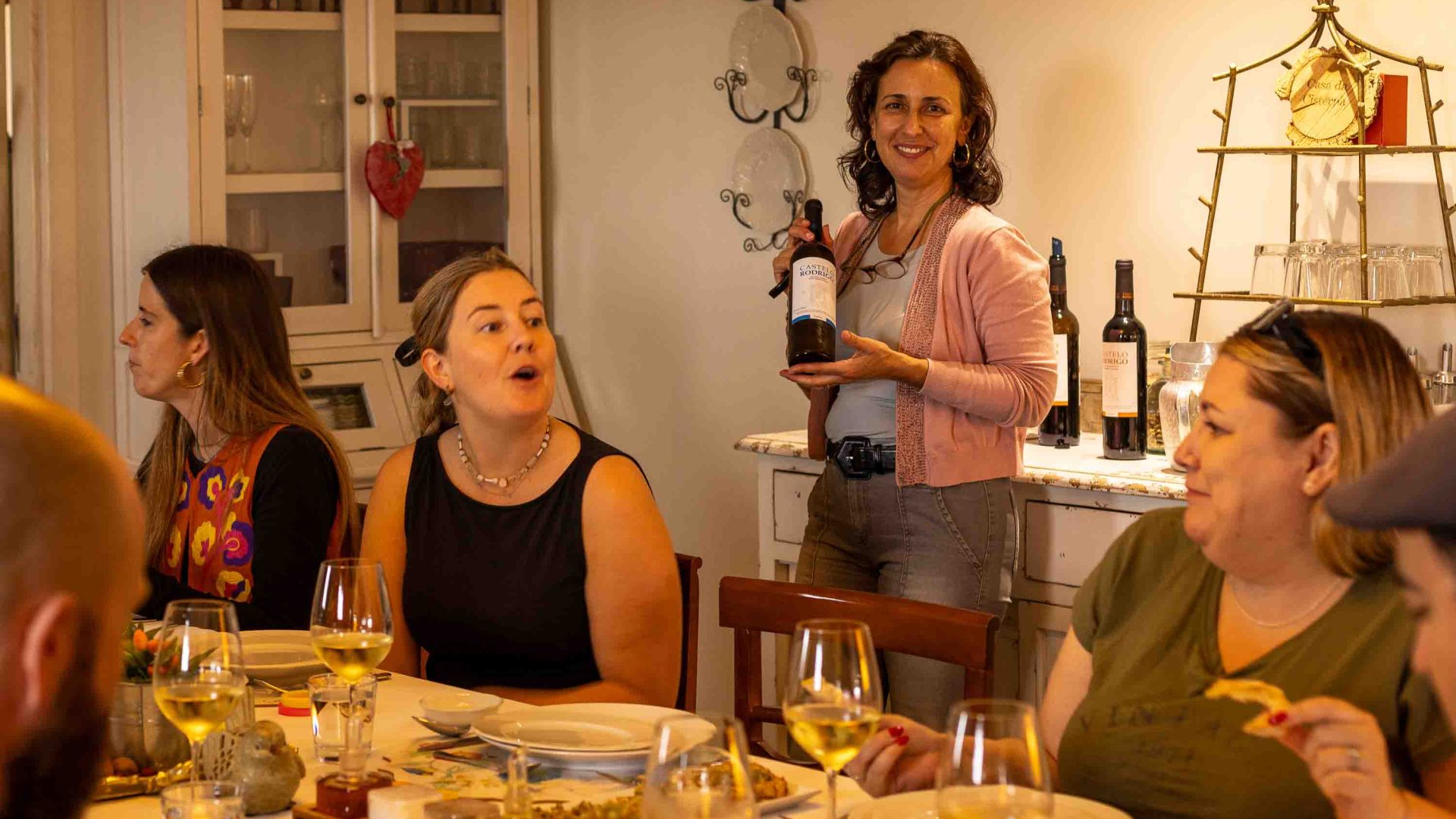 A woman holds a bottle of wine and smiles while others dine at a table.