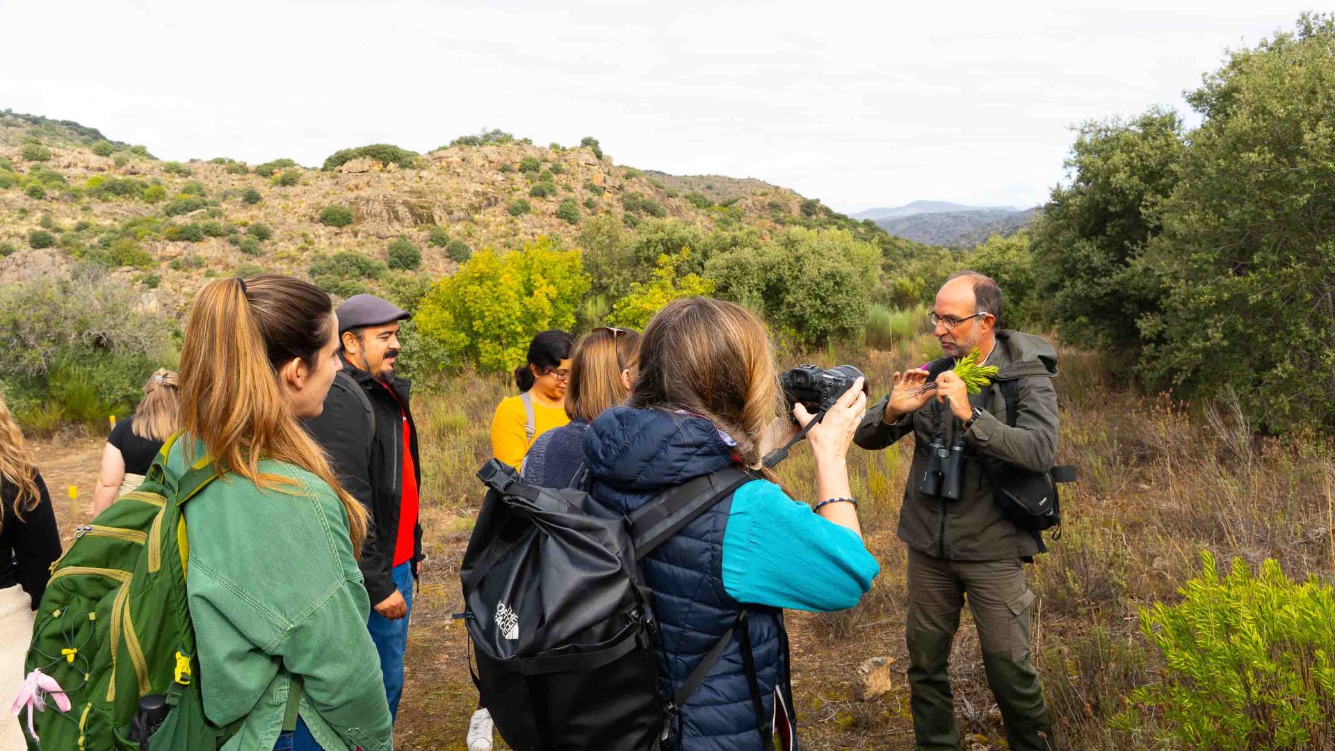 A group of people listen to a man talking. They are in a forested setting.