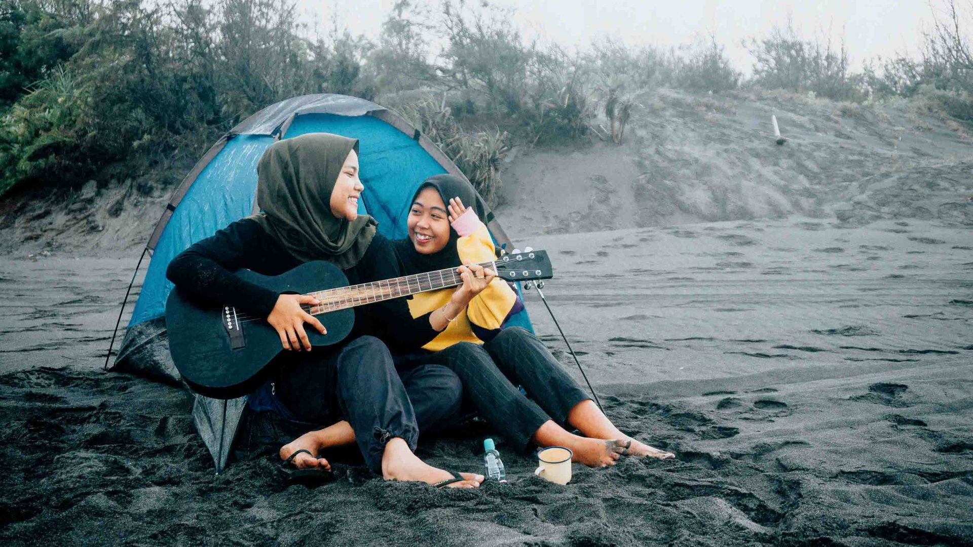 Two women in hijab's play guitar outside their tent on a beach.