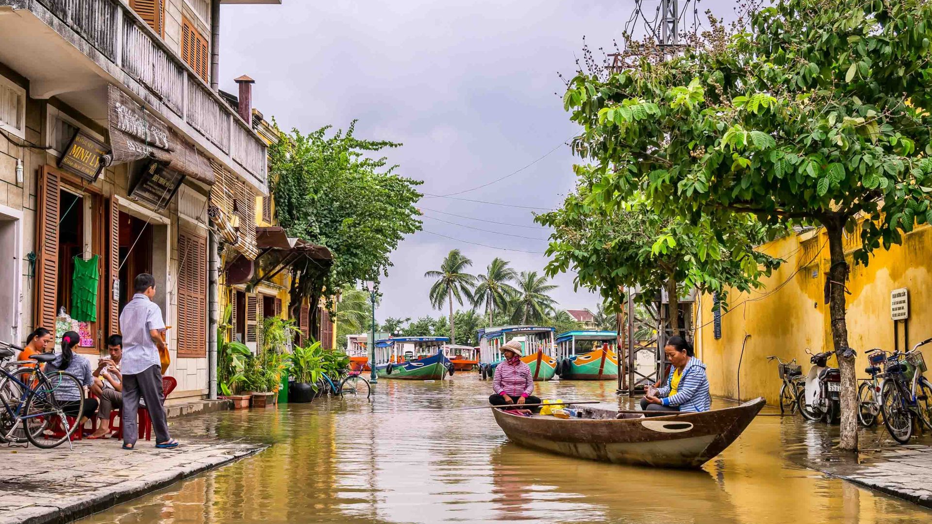 Some Vietnamese people row down a flooded street between houses while others play cards on the sidewalk.