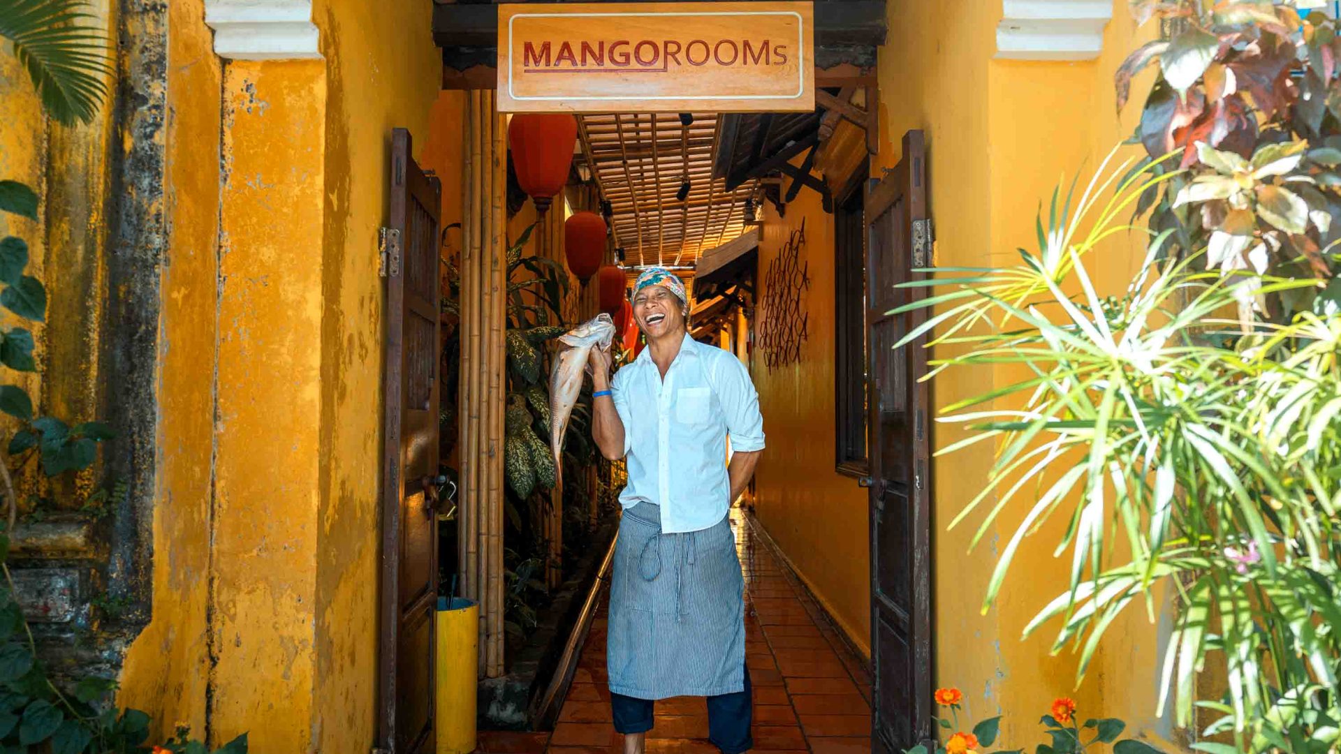 A man holds up a fish and laughs in front of a yellow building.