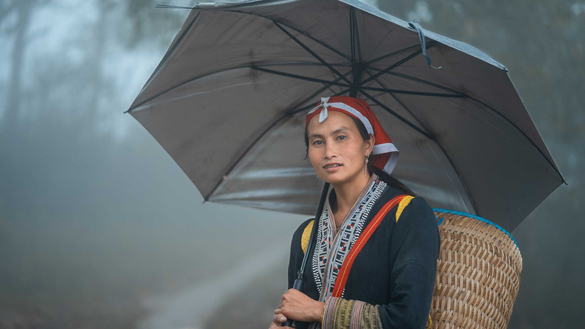 A woman in traditional clothing holds an umbrella. She has a basket on her back.