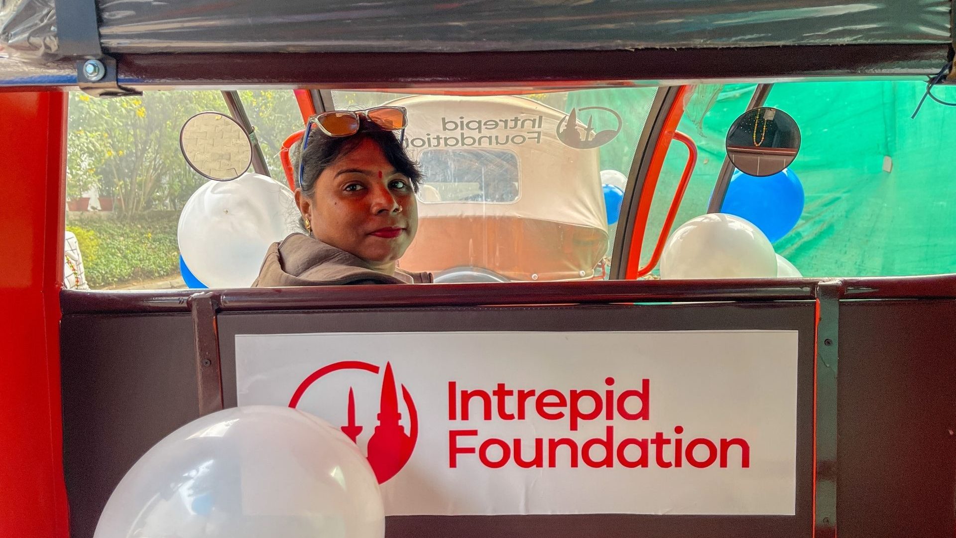 A woman at the wheel of her rickshaw looks back to the passenger seat with an Intrepid Foundation sign in the foreground
