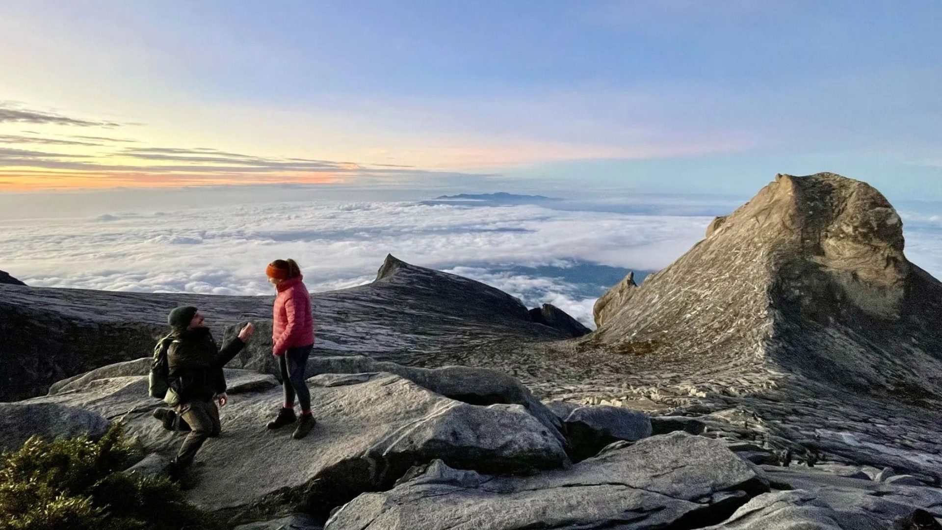 A man on one knee proposes to a woman on a mountain summit with sunrise and clouds in the background.