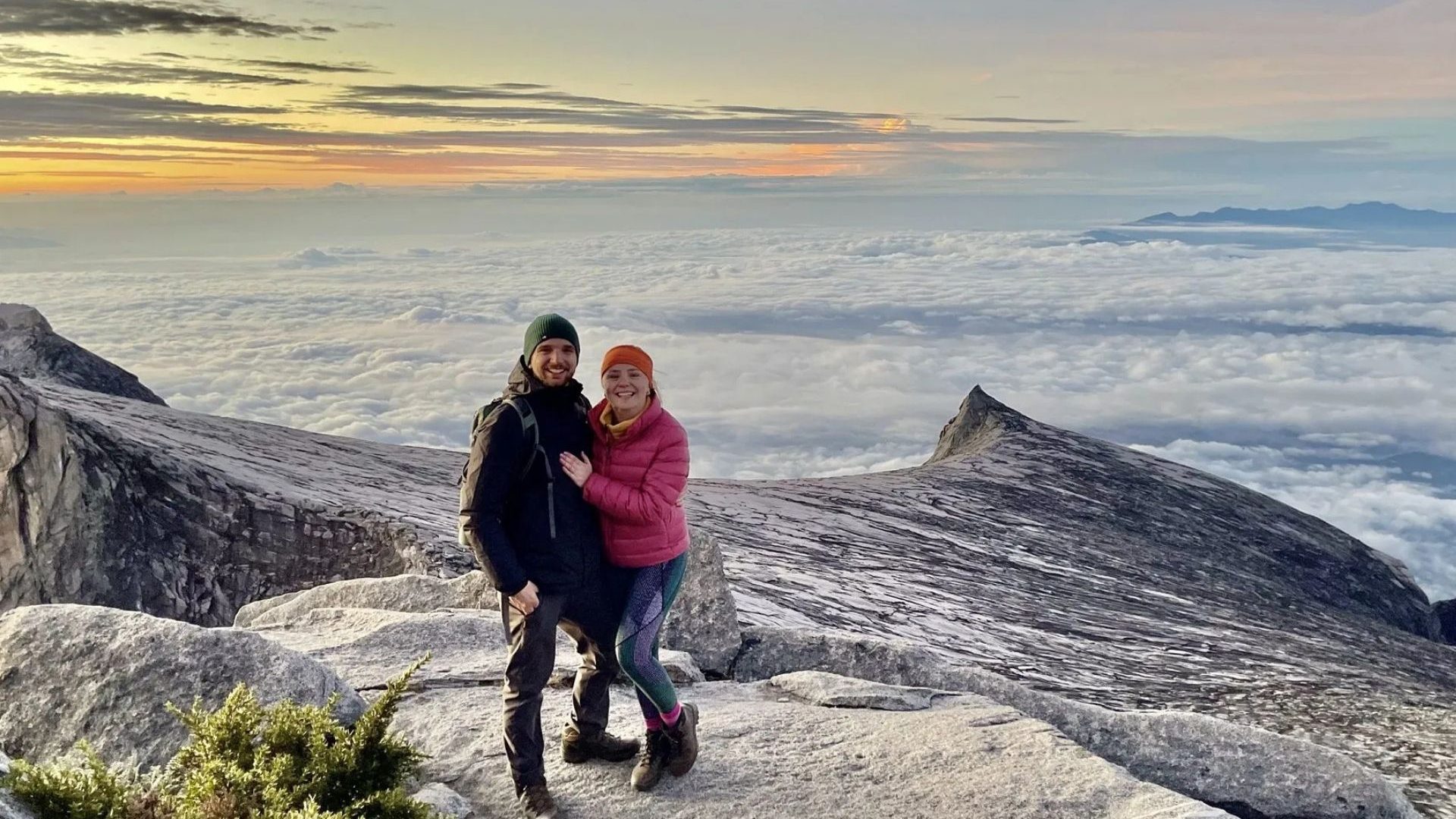 A man and woman smile from a mountain summit with sunrise and clouds in the background.