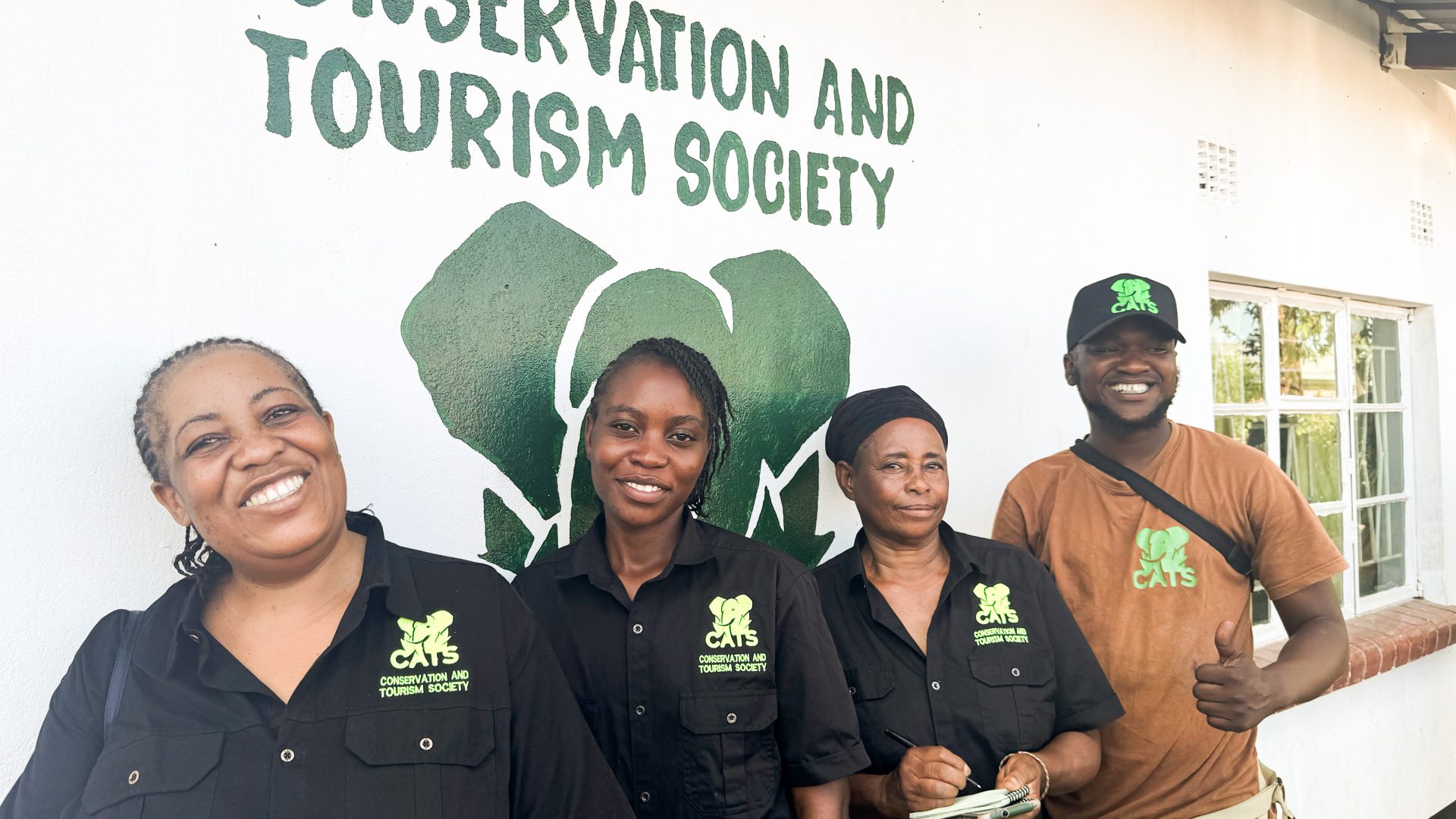 Four people stand underneath a painted sign on the wall that reads 'Conservation and Tourism Society'