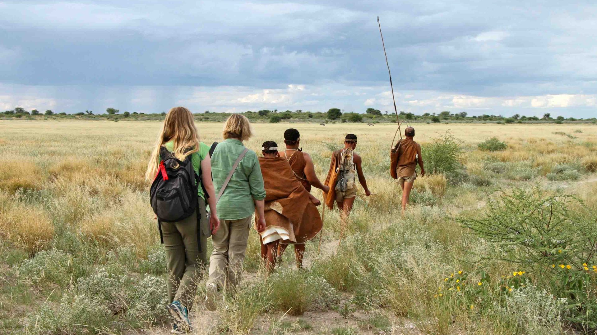Two tourists walk behind some trackers along a path through grasslands.