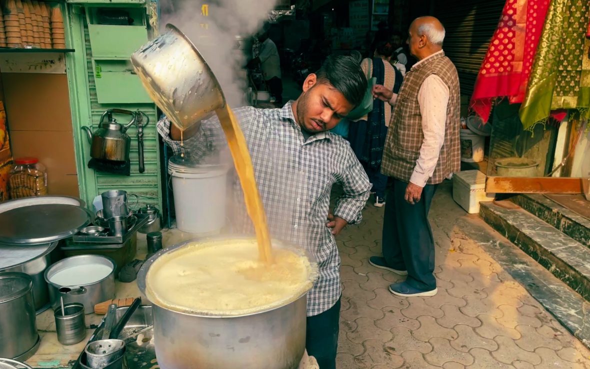 A man pours tea from a steel pan into a larger pan at a streetside masala chai stand.
