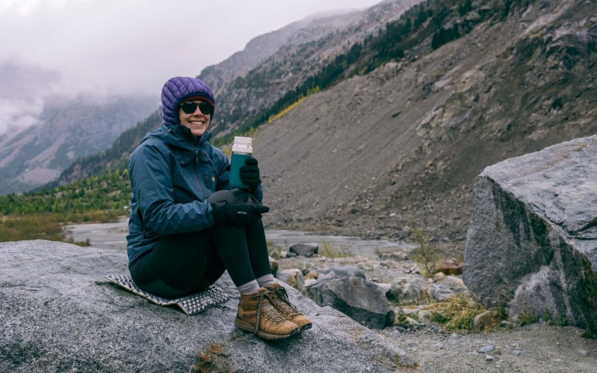 A woman wearing a blue rain jacket with a purple puffy hood sticking out smiles while sitting on a Therm-a-Rest Z-Seat atop a wet rock in the Swiss Alps.