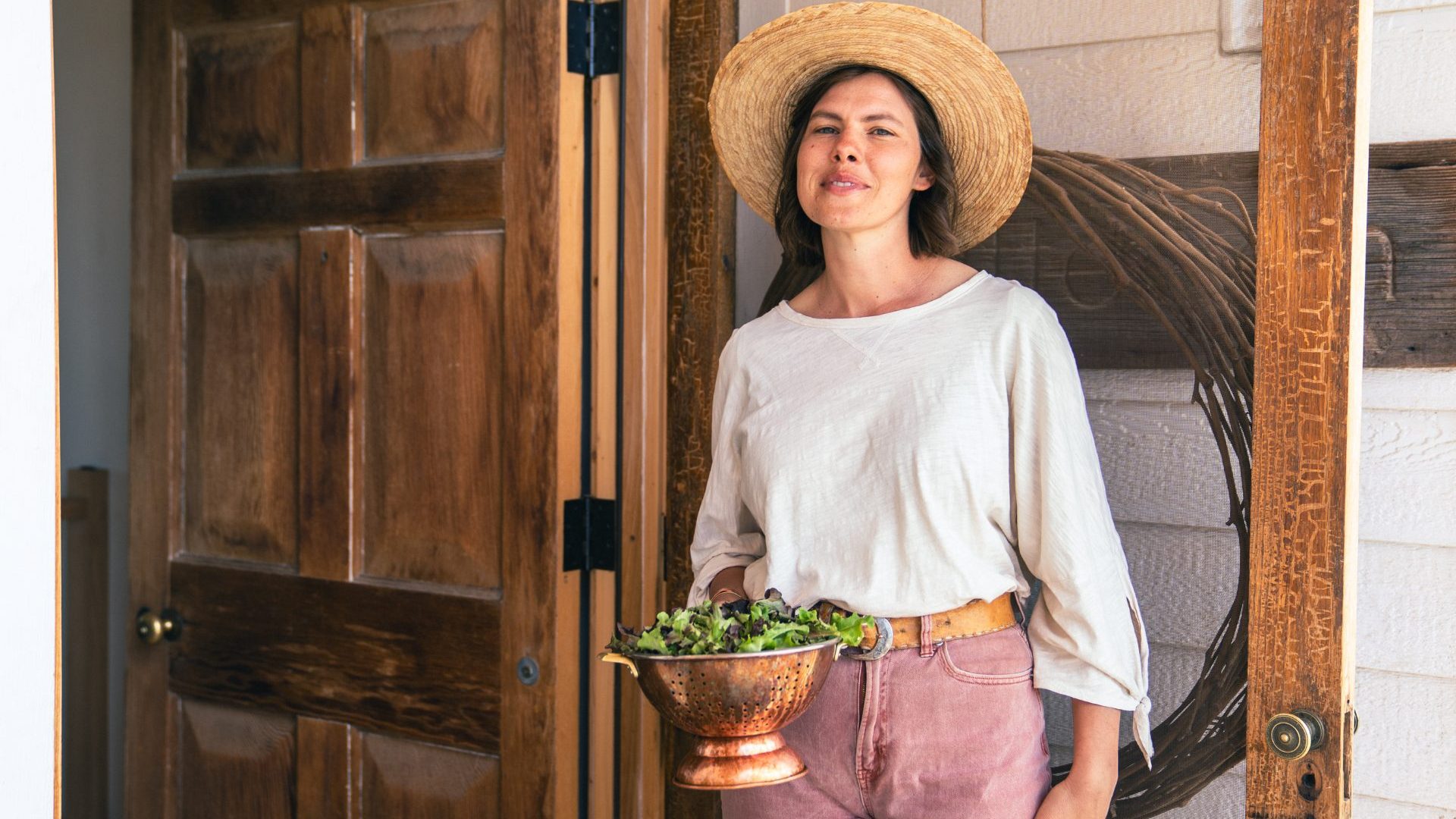Woman wearing straw hat poses for a photo holding bowl of fresh picked greens