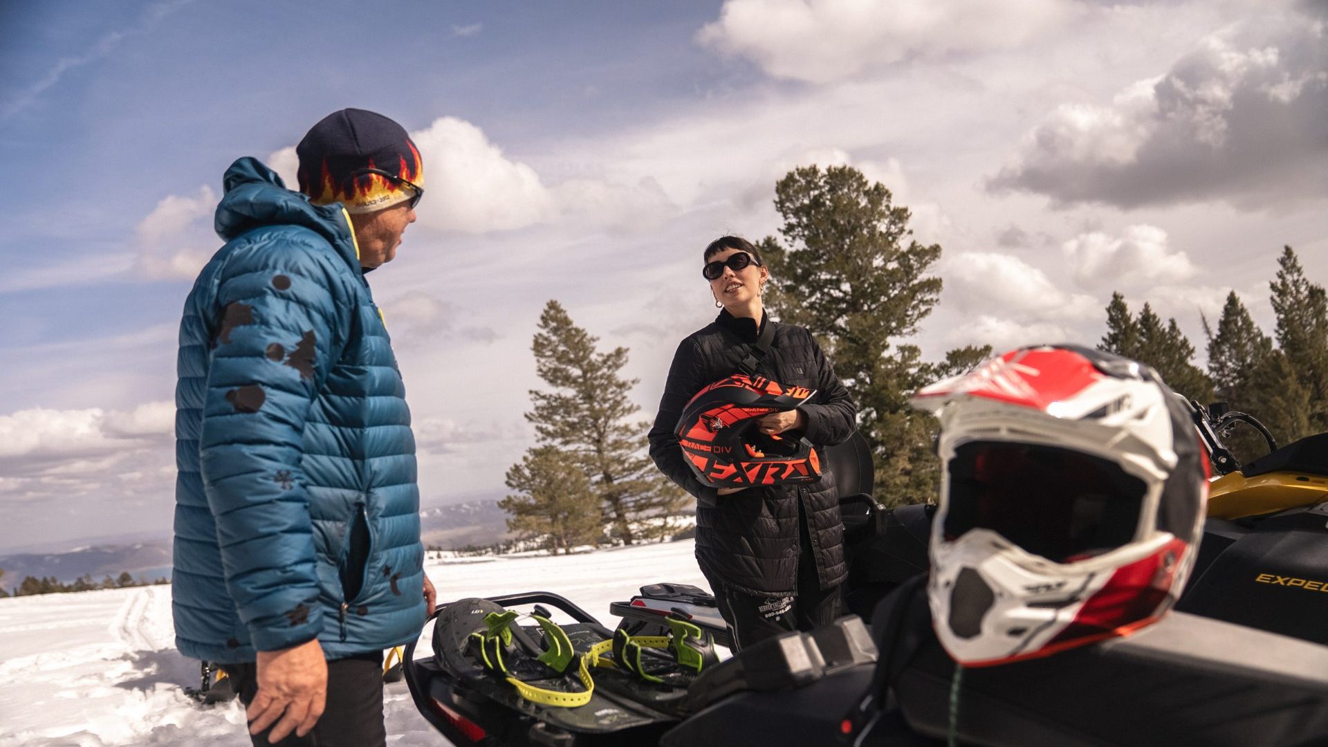 Man and woman stand in the snow next to snowmobiles