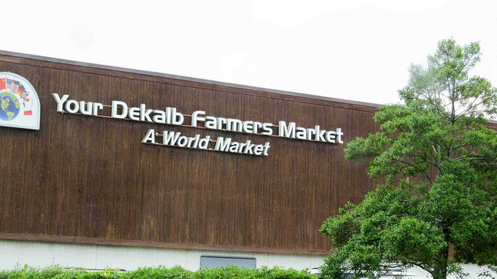 The exterior of the Dekalb Farmers Market has white letters on brown bricks and green plants with yellow flowers.