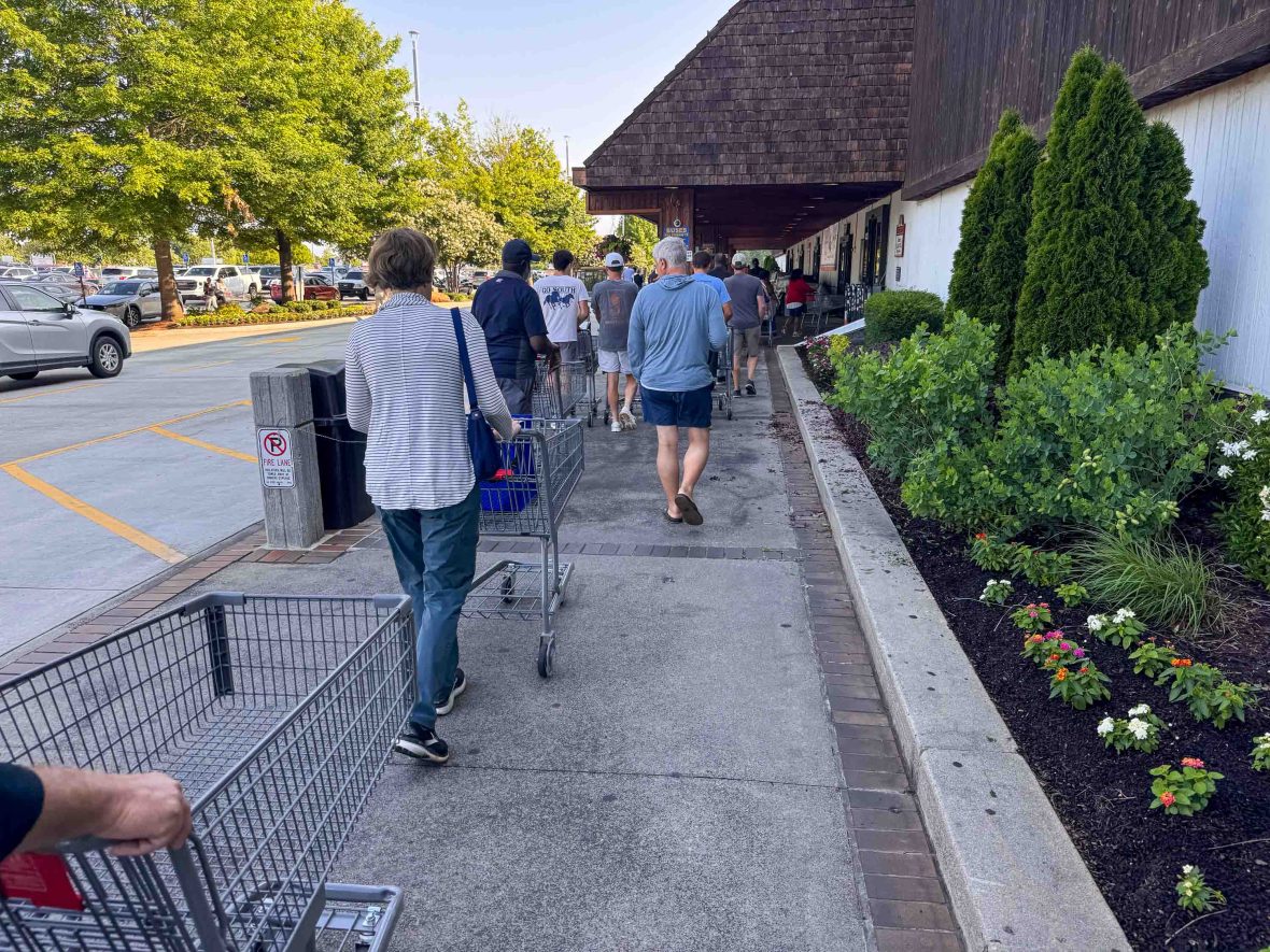 A queue of people with shopping carts waits outside the market.