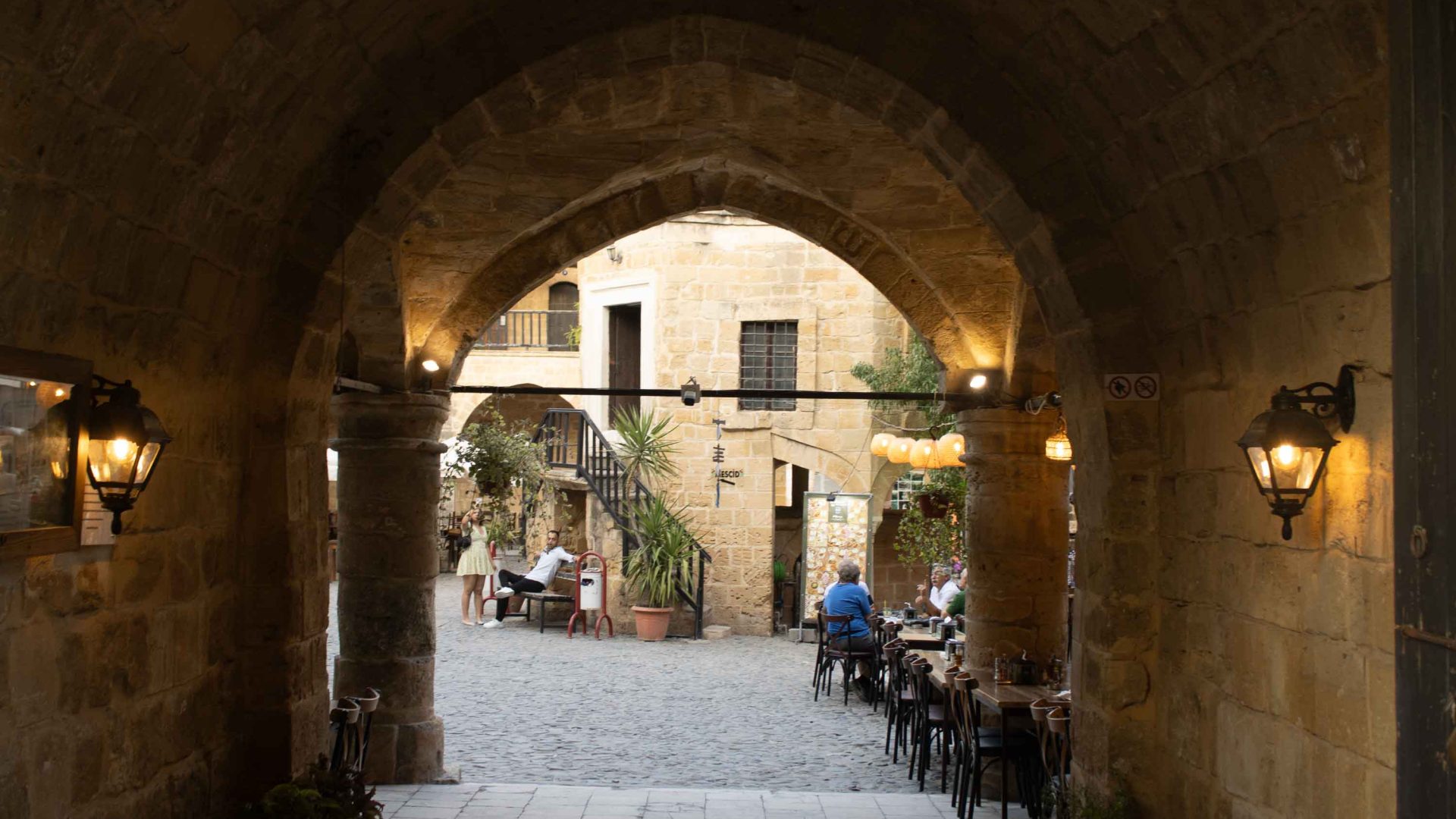 An arch leads to a plaza with dining tables and umbrellas.