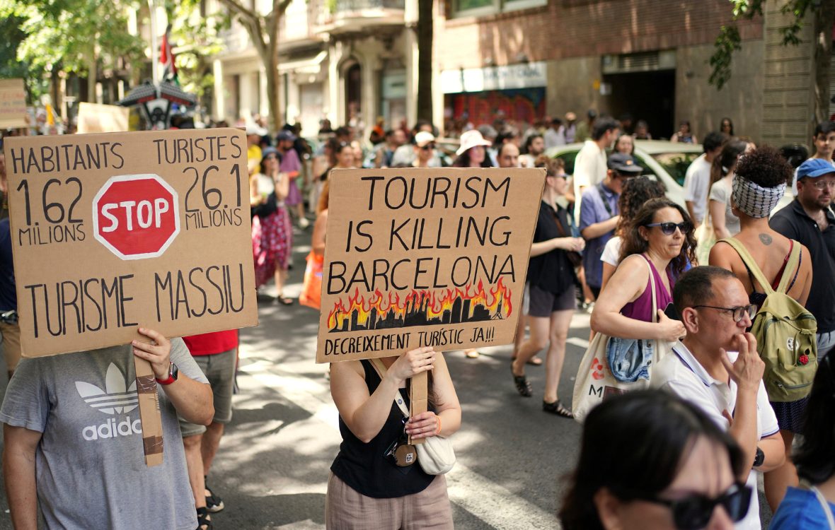 Protestors hold up placards. One says 'Tourism is killing Barcelona' and another says 'Stop' with information that Barcelona has 1.62 million inhabitants but 26.1 million tourists.
