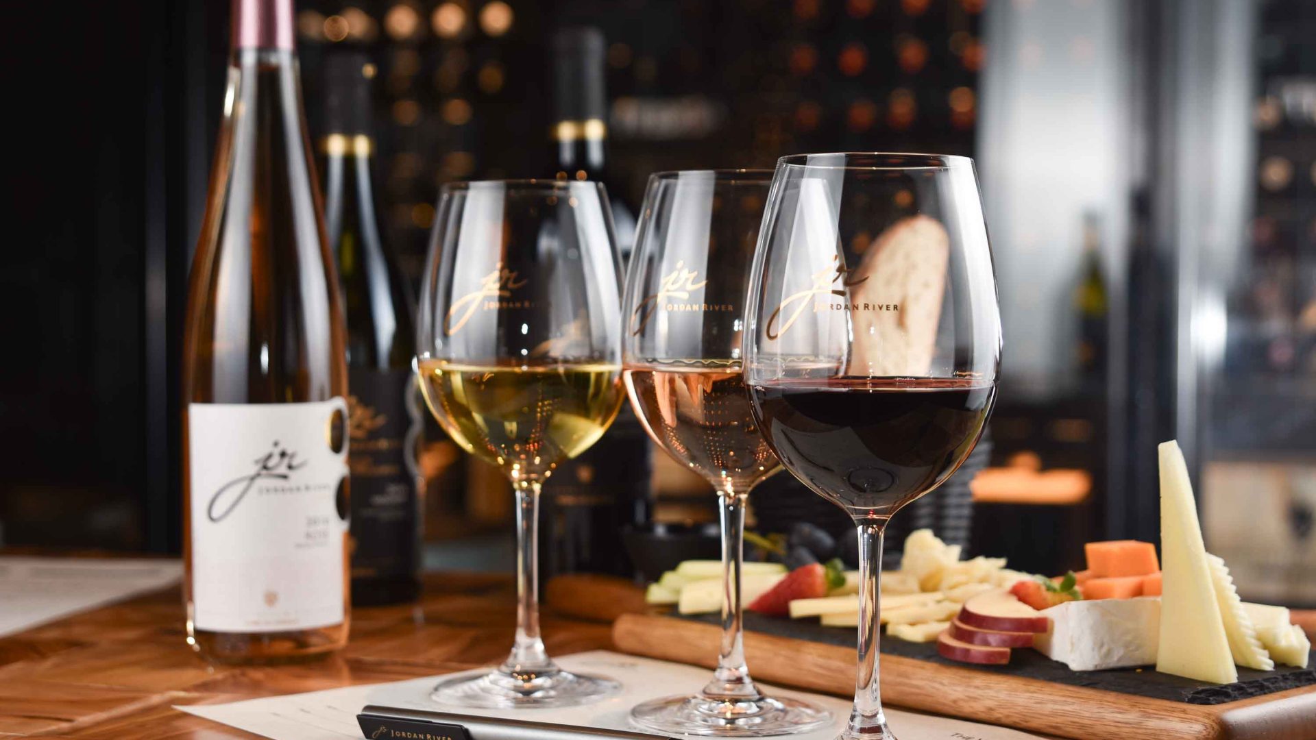 A bottle of red wine alongside three half filled glasses and a snack board.
