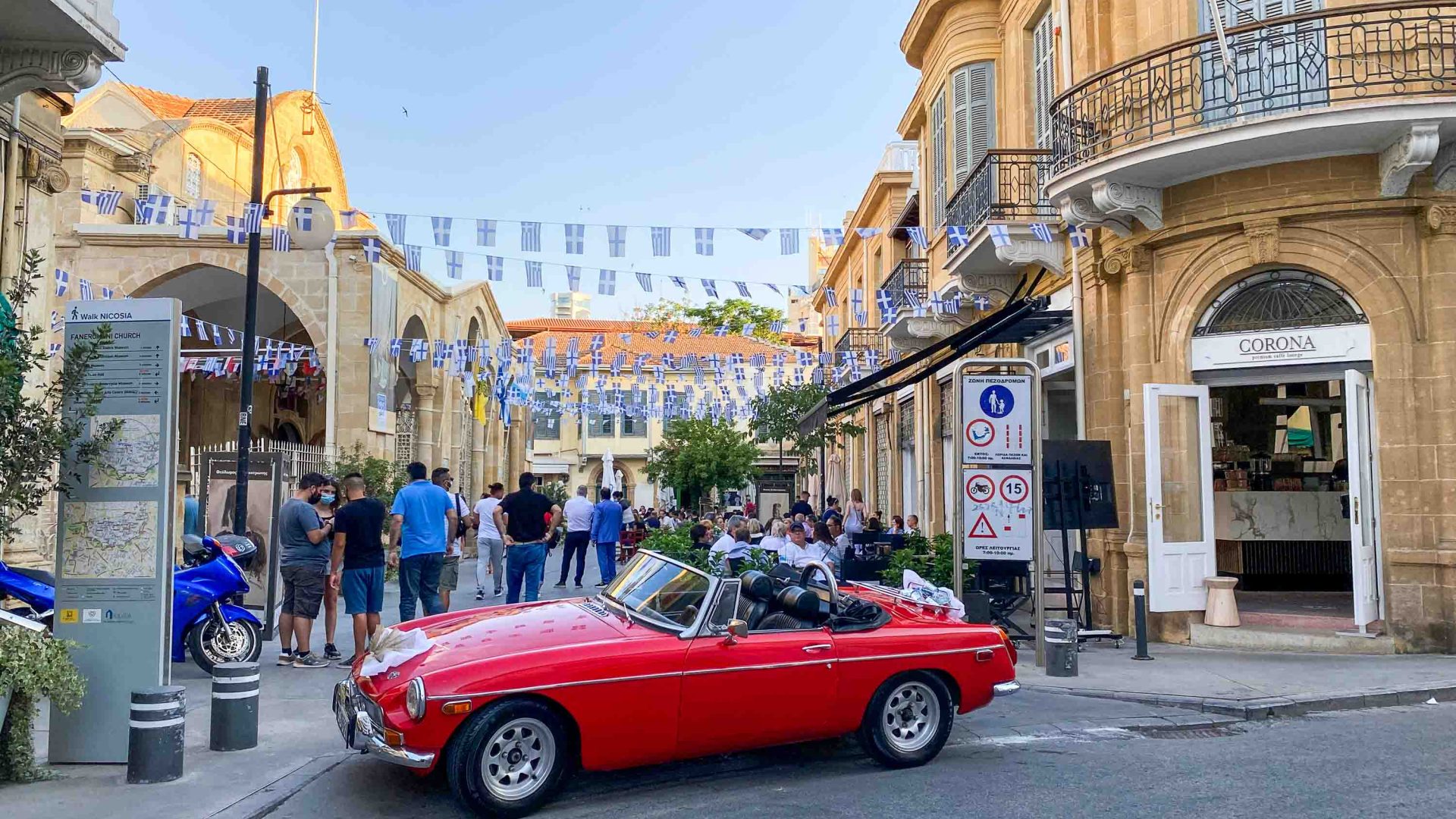 A red MG on the street, next to an event taking place with Greek flag bunting.