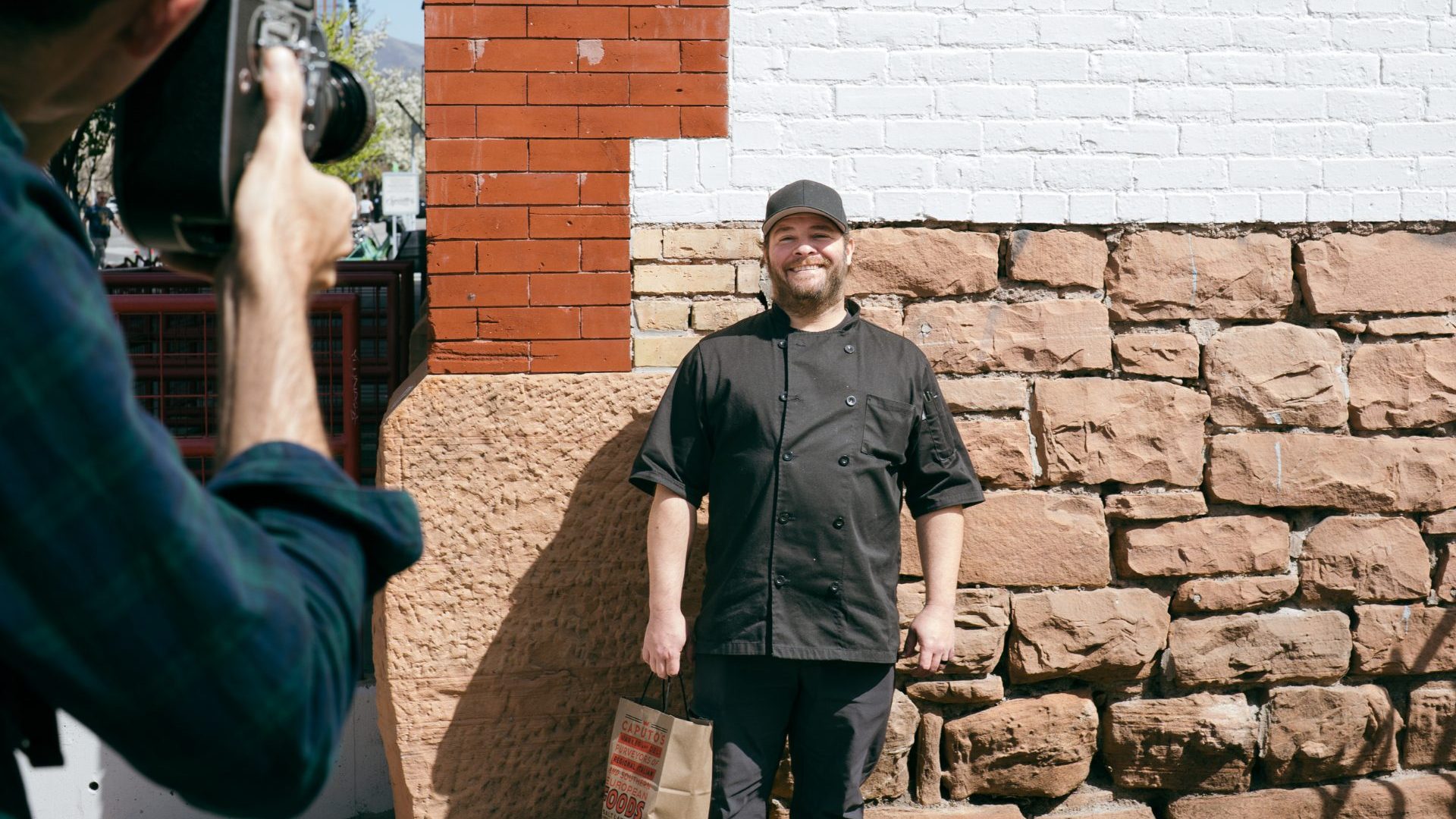Man in black chefs uniform poses for photo in front of brick door