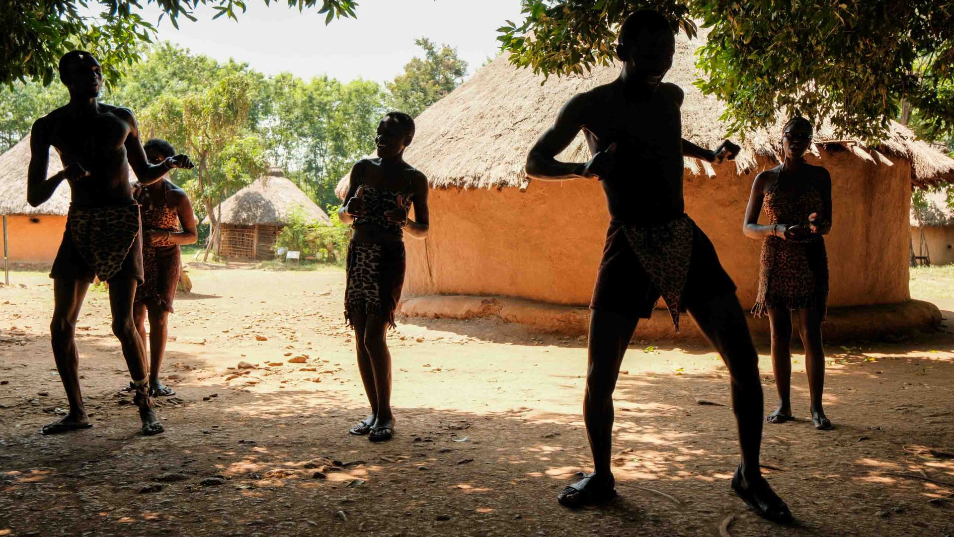 People dance in front of some mud huts.